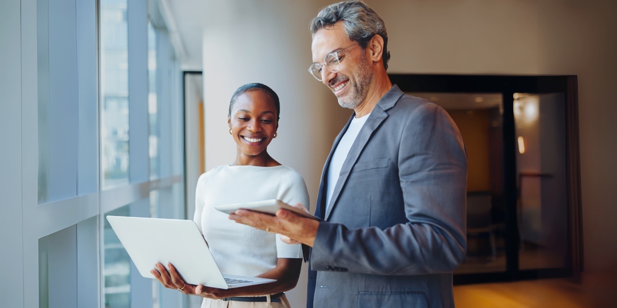 Two business professionals standing by windows, one using a laptop, one with a tablet